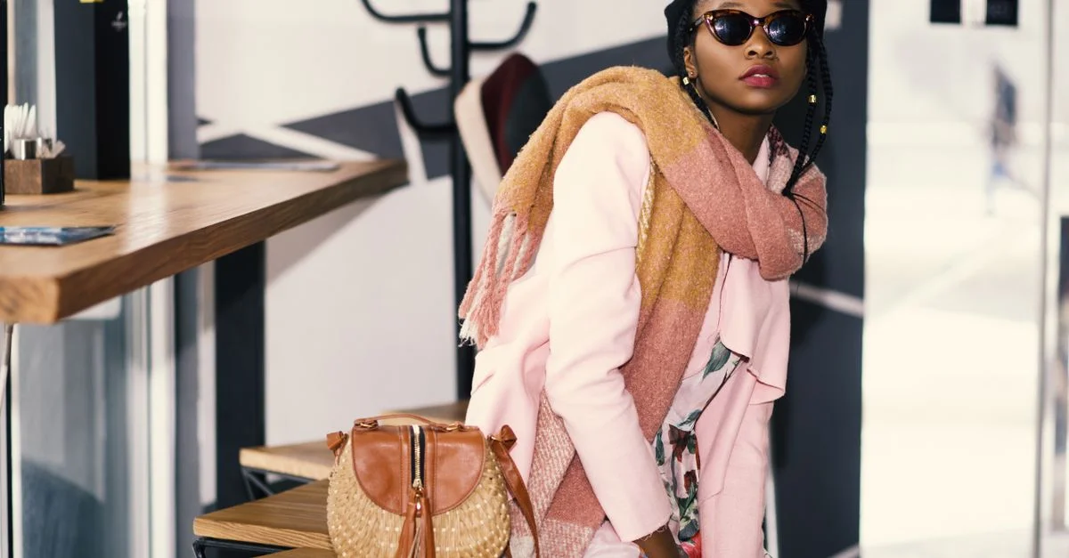 Femme africaine chic avec des lunettes de soleil et une écharpe assise à l'intérieur à côté d'un sac élégant.