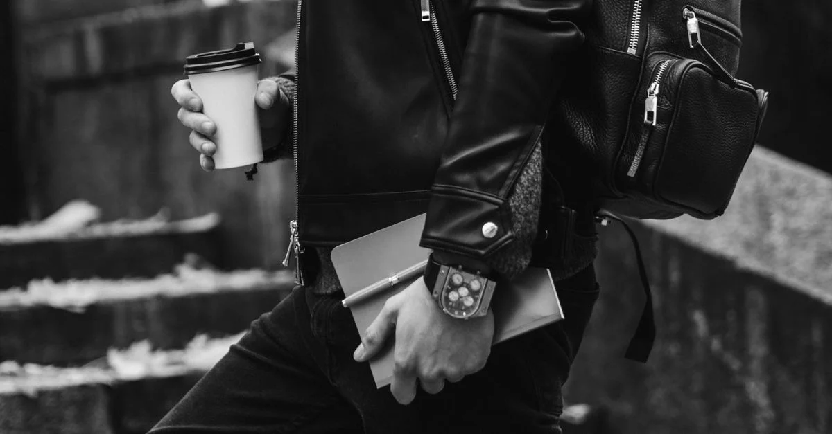 Photo en noir et blanc d'une personne tenant une tasse de café, portant une veste en cuir et un sac à dos.