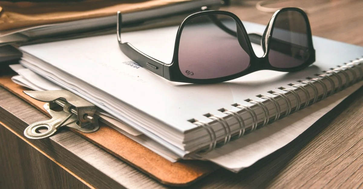 Une paire de lunettes de soleil reposant sur un carnet à spirales et un porte-bloc sur un bureau en bois.