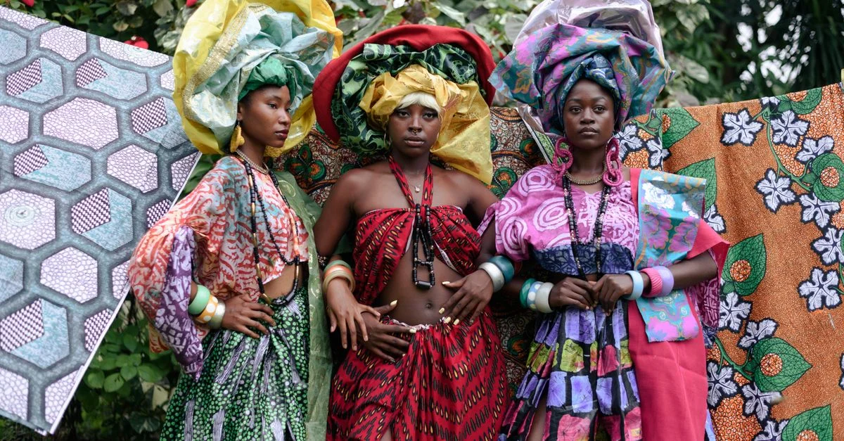 Trois femmes vêtues de tenues nigérianes colorées et coiffées de foulards, posent avec assurance en extérieur.