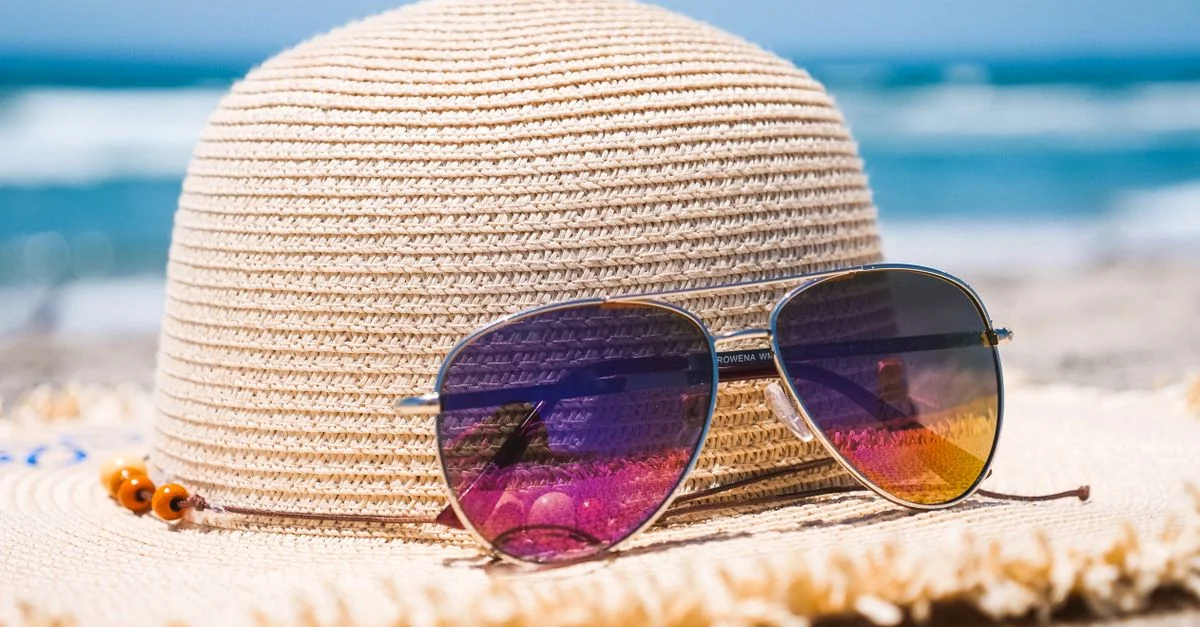 Gros plan sur un chapeau en paille et des lunettes de soleil sur une plage de sable avec des vagues océaniques en arrière-plan.